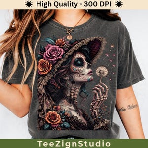 Könnte beinhalten: Dunkelgraues T-Shirt mit einer detaillierten Illustration einer Sugar-Skull-Frau, die mit Blumen geschmückt ist und einen Löwenzahn hält. Das Design weist komplizierte Details und lebendige Farben auf, mit dem Text "TeeZignStudio" unten.