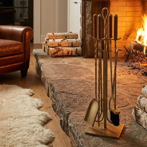 May include: A fireplace scene featuring a set of gold-toned fireplace tools, including a shovel, brush, and poker. A burning fire is visible in the background, with stacked birch logs nearby. A brown leather armchair and a white sheepskin rug are also in the scene.