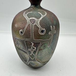 Puede incluir: Un jarrón decorativo con una superficie texturizada y un borde azul oscuro. El jarrón presenta un diseño tribal de color plateado. El cuerpo del jarrón tiene una mezcla de tonos marrones, verdes y azules.