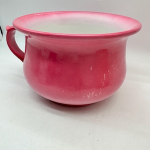 Op de afbeelding: Een kamerpot in vintage-stijl met een felroze buitenkant met een glanzende afwerking en een witte binnenkant. De kom heeft een brede, uitlopende rand en een enkele gebogen handgreep aan de zijkant, met lichte gebruikssporen en witte krassen.