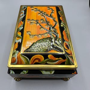 May include: Rectangular black box with a gold trim and feet. The lid features a detailed painting of a bonsai tree against an orange background. The sides are decorated with colorful floral patterns.