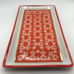 May include: Rectangular ceramic tray with a white border and a red rim. The center features a repeating pattern of white floral designs on an orange background, with gold accents.