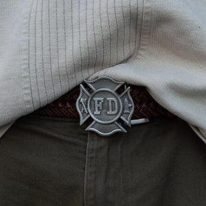 May include: A silver belt buckle with a cross and the letters "FD" in the center. The buckle is attached to a brown leather belt.