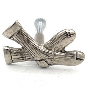 May include: A silver-toned metal cabinet knob in the shape of a pair of socks. The knob has a textured surface and a screw for attachment. The socks are depicted in a crossed arrangement.