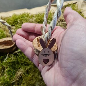 May include: A small wooden bunny basket with a heart cutout and a rope handle. The basket is made from a halved walnut shell and is sitting on a bed of green moss.
