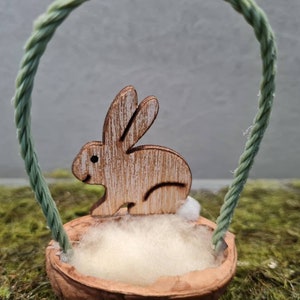 May include: A wooden bunny figurine sits inside a small, natural walnut shell with a green rope handle. The bunny is painted white with brown accents and has a fluffy white cotton lining inside the shell.