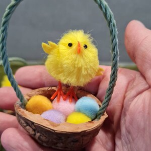 May include: A small yellow chick with red legs stands on top of a walnut shell basket filled with colorful pom-pom Easter eggs. The basket has a green rope handle.