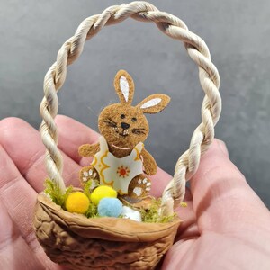 May include: A small brown bunny rabbit figurine sits in a walnut shell basket with a rope handle. The basket is filled with moss and colorful Easter eggs.