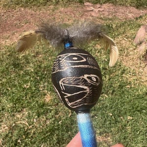 May include: A round, dark brown rattle featuring etched animal designs on its surface. The top is adorned with grey and tan feathers attached with blue thread, while the handle is wrapped in a blue and white gradient fabric covering.