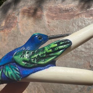 May include: A hair clip featuring a sculpted hummingbird with a vibrant blue head and body, accented by textured green wings and foliage details. The bird is attached to a smooth, off-white, two-pronged hair accessory with a long, thin black beak.