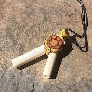 May include: A handcrafted pendant necklace featuring a turtle design. The turtle pendant has a brown and tan shell with green eyes, and is attached to two off-white cylindrical pieces. The necklace has a black cord.