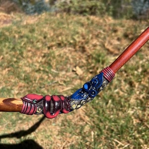 May include: A wooden wand featuring a sculpted red snake coiled around the shaft, transitioning into a blue floral ornament with metallic accents. The handle is smooth, polished brown wood, while the decorative section displays intricate textures.