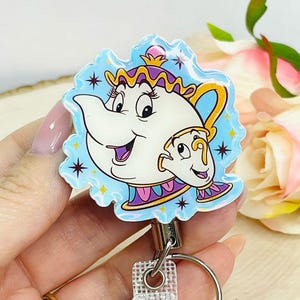 May include: A badge reel featuring a cartoon image of Mrs. Potts and Chip from Beauty and the Beast. The design is set against a light blue background with star accents. The reel has a clear plastic clip and a silver-colored metal ring.