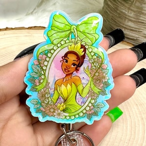 May include: A badge reel featuring a cartoon image of a princess in a green dress, framed by a teal border and a green bow. The reel has a clear plastic strap and a metal clip for attaching to clothing.