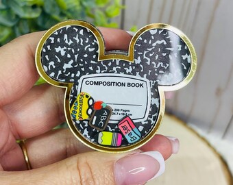 May include: A Mickey Mouse-shaped badge reel featuring a black and white speckled composition book pattern. The center displays a white label with the text COMPOSITION BOOK, 200 Pages, and 24.7 x 19.0 cm, surrounded by miniature school-themed charms.