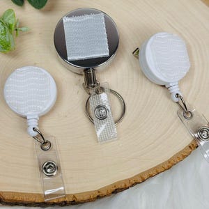 May include: Three white and silver retractable badge holders with clear plastic straps and metal key rings. The badge reels have a white, textured surface for attaching a badge. The background is a light-colored wooden surface.