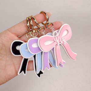 May include: A set of four layered acrylic bow keychains in black, light blue, lavender, and pink. Each bow features a white outline and a gold-toned metal lobster clasp. The pink bow has the name Liz written in white script lettering on the front.
