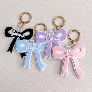May include: Four bow-shaped keychains with gold-toned metal rings. Each keychain features a different color: black, light blue, lavender, and pink. The names Gemma, Isabel, Connie, and Liz are written in white script lettering across the center.