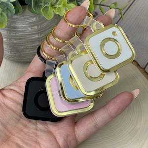 May include: A set of five keychains featuring a square design with a gold rim and a white circle in the centre. The keychains are in different colours: black, pink, light blue, beige, and white.