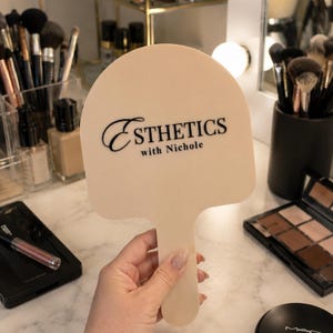 May include: A beige, arch-shaped handheld mirror featuring the black printed text Esthetics with Nichole centered on the front. The mirror has a smooth, solid-colored surface and a rounded handle, providing a clear view of the brand name.