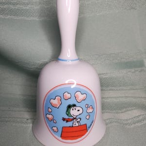 May include: A white ceramic hand bell featuring a circular graphic of Snoopy wearing a green aviator cap and red scarf, sitting on a red doghouse against a blue sky with pink heart-shaped clouds. The bell has a light blue ring at the handle base.