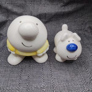 May include: A pair of white ceramic salt and pepper shakers. The larger shaker is round with a yellow shirt and a smiling face. The smaller shaker is shaped like a dog with a blue nose, pointed ears, and a smiling face, featuring two holes on top.