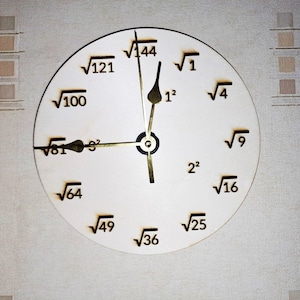 May include: A white clock face with black numbers representing square roots and exponents. The clock hands are gold and point to 12 and 3.