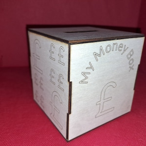 May include: A white wooden money box with a slot on the top. The box has a design of pound sterling symbols on the front and the text "My Money Box" on the side.