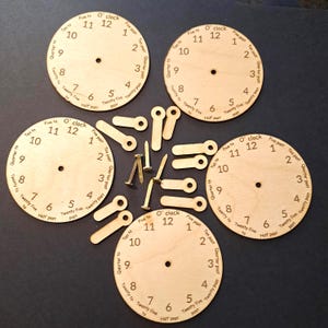 May include: Wooden clock faces with numbers and labels, plus wooden clock hands and brass nails. The clock faces are light brown with black numbers and text. The hands are light brown. The items are arranged on a dark surface.