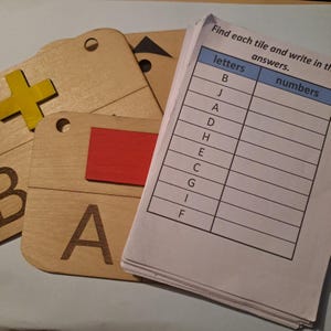 May include: A wooden educational game with letter and number tiles. The tiles are light brown with black letters and a yellow plus sign. A worksheet with the text "Find each tile and write in the answers" is also present.