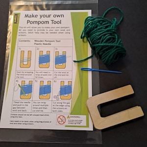 May include: A craft kit for making pompoms. The kit includes a wooden pompom tool, a plastic needle, and green yarn. The instructions are printed on a white and green card. The text on the card reads "Make your own Pompom Tool."