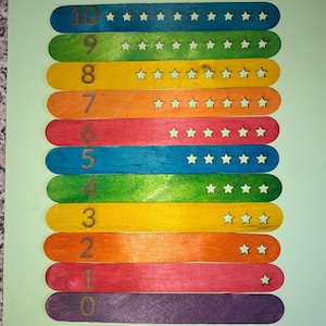 May include: A set of ten colourful wooden popsicle sticks with numbers 1 through 10 printed on them. Each stick has a corresponding number of white stars printed on it.