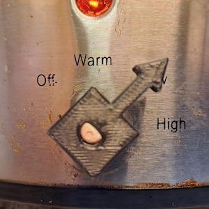 May include: A silver appliance with a black knob and a red light. The knob has a black arrow pointing to the right and the words "Off", "Warm", and "High" printed on the appliance.