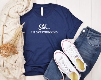 May include: A navy blue t-shirt with white text that reads "Shh... I'm overthinking". The shirt is laid flat on a white wooden surface with a white knitted scarf, dried flowers, blue jeans, and white sneakers.