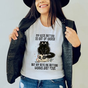 May include: A white t-shirt featuring a black cat perched on a skull. The text on the shirt reads "My nice button is out of order but my bite me button works just fine."