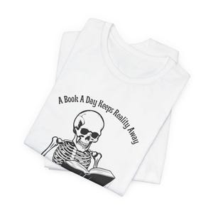 May include: White t-shirt with a black skeleton reading a book. The text on the shirt reads "A Book A Day Keeps Reality Away."