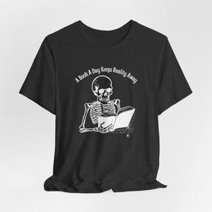 May include: A dark heather grey t-shirt with a white skeleton reading a book and holding a cup. The text "A Book A Day Keeps Reality Away" is printed on the shirt.