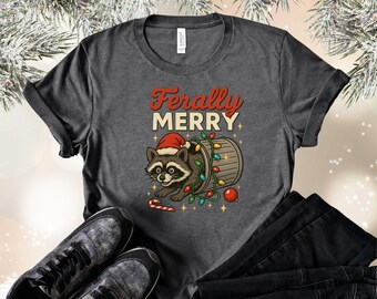 May include: Dark grey t-shirt with a cartoon raccoon in a Santa hat, emerging from a barrel decorated with Christmas lights. The text "Ferally Merry" is in red and white. A candy cane and baubles are also visible.
