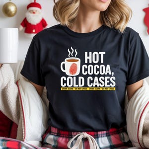 May include: Black t-shirt with the text "HOT COCOA, COLD CASES" in white. The design includes a steaming mug of cocoa with a red fingerprint. The shirt also has the text "CRIME SCENE - DO NOT CROSS" in yellow. A white mug is held in the person's hand.