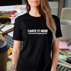 May include: Black crew neck t-shirt with the text "I HATE IT HERE but at least I'm funny about it." The text is in white. The shirt is worn by a person in an office setting.