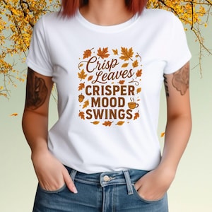 May include: White t-shirt with a fall-themed graphic. The design features the words "Crisp Leaves, Crisper Mood Swings" in a brown, stylized font, surrounded by autumn leaves, an acorn, and a coffee cup.