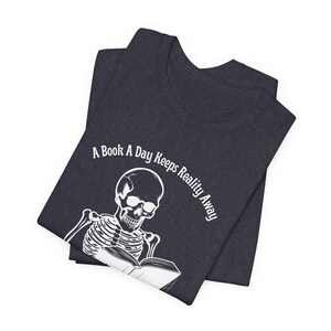 May include: A dark blue t-shirt with a white skeleton reading a book. The text on the shirt reads "A Book A Day Keeps Reality Away".