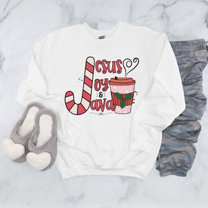 Peut inclure: Sweat-shirt blanc avec l'inscription "Jesus, Joy & Java" en rouge et rose. Le motif comprend un sucre d'orge et une tasse de café avec du houx. Des pantoufles grises en forme de cœur et un pantalon camouflage sont également visibles.