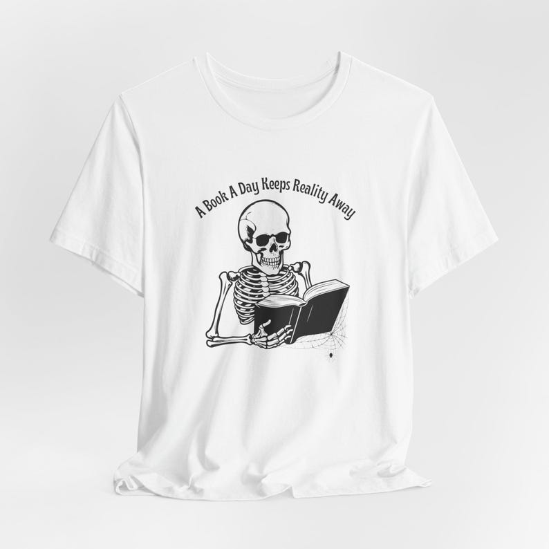 May include: White t-shirt with a black and white illustration of a skeleton reading a book. The text "A Book A Day Keeps Reality Away" is printed on the shirt.