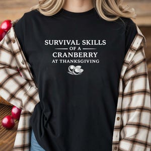 May include: Black t-shirt with the text "SURVIVAL SKILLS of a CRANBERRY at THANKSGIVING" in white. A cranberry graphic is below the text. The shirt is worn with a plaid button-down shirt.