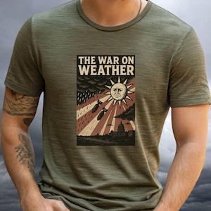 Peut inclure: T-shirt vert olive avec un motif graphique portant l'inscription "THE WAR ON WEATHER". Le motif comprend un soleil renfrogné, des bombes qui tombent et une silhouette de bâtiment. Le t-shirt a des manches courtes et un col rond.