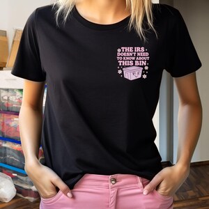 May include: Black t-shirt with the text "THE IRS doesn't need to know about THIS BIN." in pink. The design includes a cartoon bin and snowflakes. The shirt is a crew neck and is being worn.