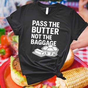 May include: Black t-shirt with the text "PASS THE BUTTER NOT THE BAGGAGE" in white. The graphic includes a stick of butter and a basket of rolls. The shirt is knotted at the waist and is on a red tray with food.