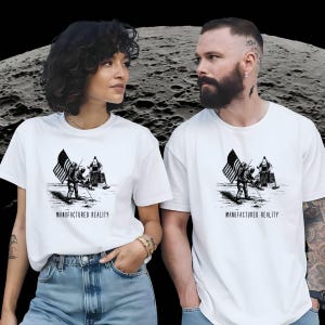 May include: Two white t-shirts with a black and white illustration of an astronaut planting a flag on the moon. The text "MANUFACTURED REALITY" is printed below the illustration.
