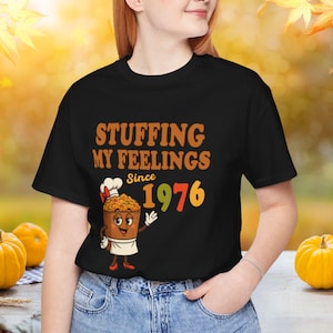 May include: Black t-shirt with the text "STUFFING MY FEELINGS Since 1976". The shirt features a cartoon image of a bowl of stuffing wearing a chef's hat and apron. The numbers "1976" are in orange and green.
